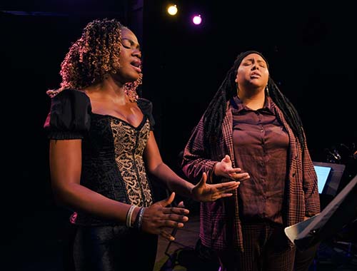 Two women passionately singing together in a dimly lit room filled with music.