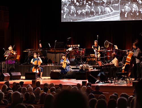 Image of musicians playing on stage at a concert, with a large screen showing dynamic visuals in the background.